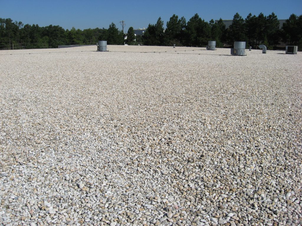 Flat commercial roofing surface covered with gravel, showcasing HVAC units and surrounding trees, relevant to roofing services and insurance claims.