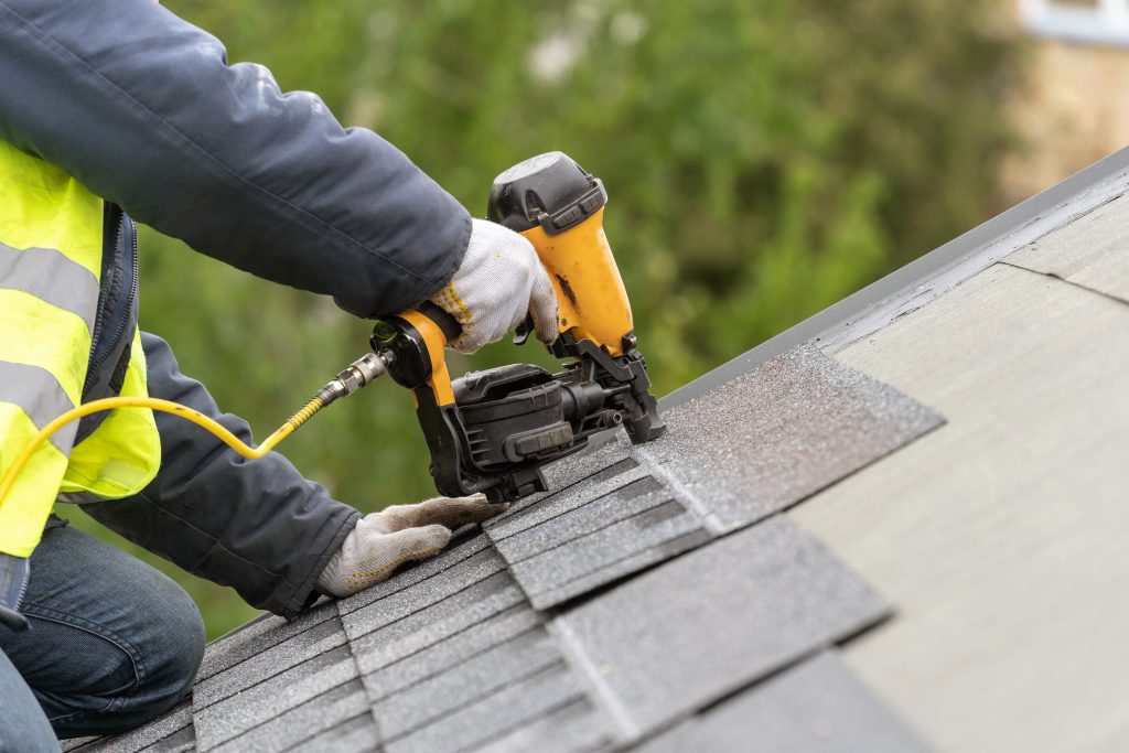 Roofer using a nail gun to install asphalt shingles on a residential roof, emphasizing roof repair and maintenance.