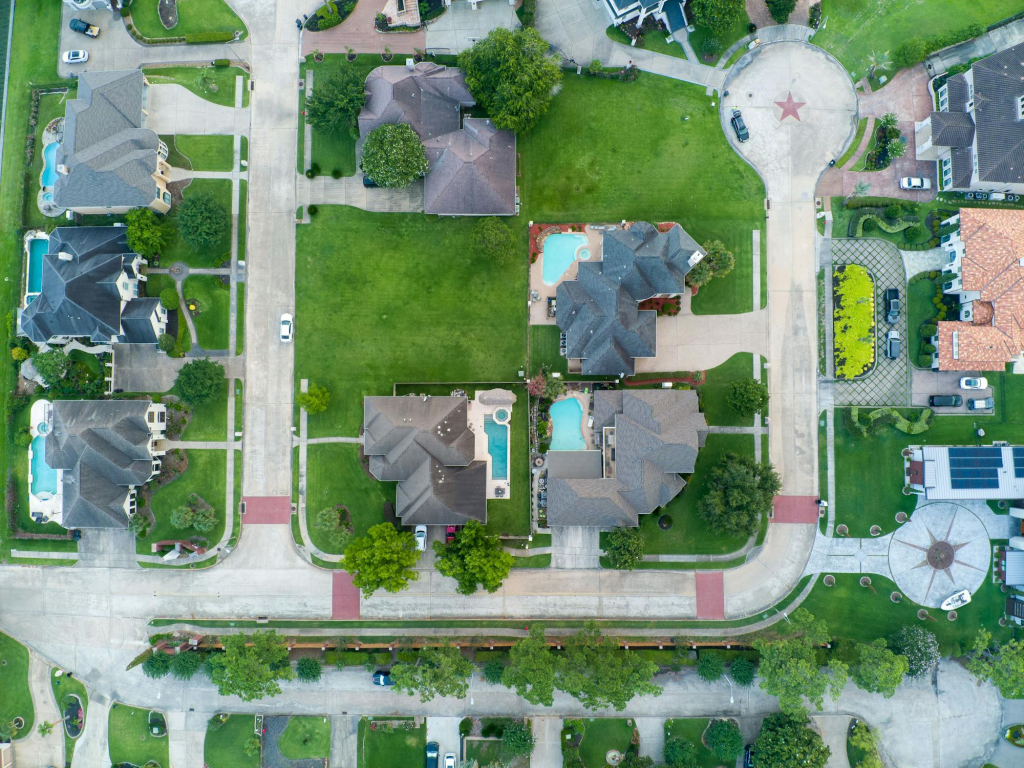 Aerial view of residential neighborhood with various houses, green lawns, and swimming pools, illustrating properties potentially affected by hail damage, relevant to roof inspection and replacement discussions.