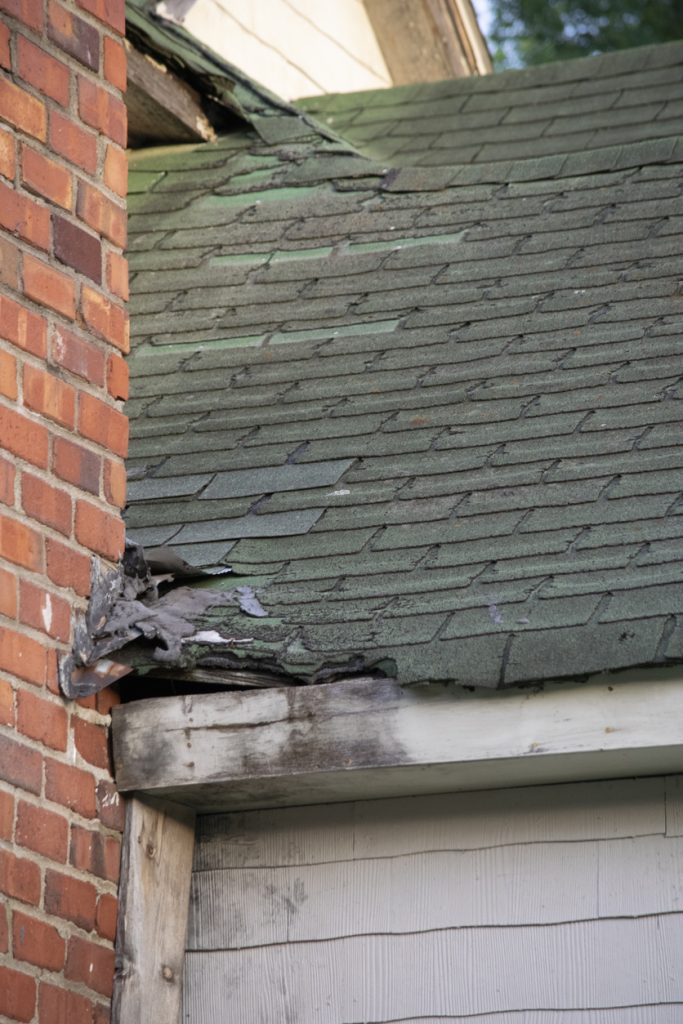 Damaged residential roof showing missing shingles and deterioration near a brick wall, highlighting the importance of professional roofing inspections and repairs for insurance claims.
