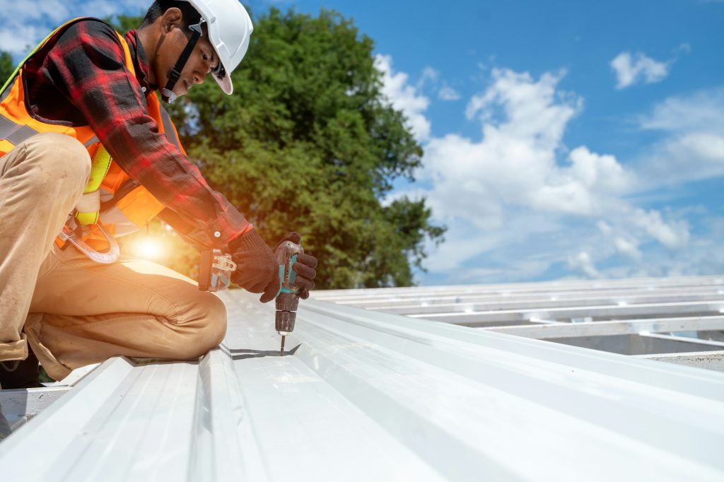 Roofer installing metal roofing with electric drill, wearing safety gear, on a bright day in Asheville, NC, highlighting Grier Roofing's expertise in durable roofing solutions.