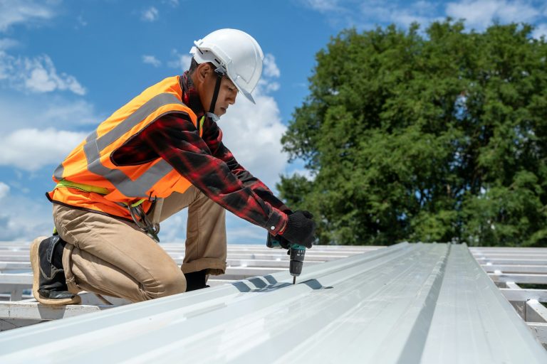 Roofer in safety gear installing metal roofing on commercial building under clear blue sky.