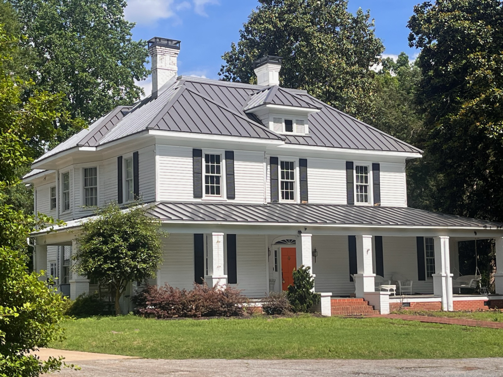 Historic white house with a metal roof, black shutters, and a welcoming porch, showcasing its well-maintained exterior in a residential setting, relevant to roof damage insurance claims.