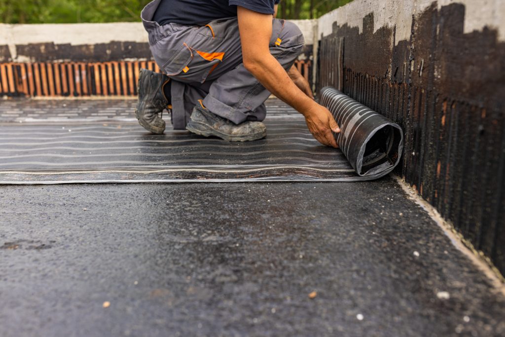 Worker installing modified bitumen roofing, laying vapor barrier on flat commercial roof, emphasizing durability and maintenance benefits.