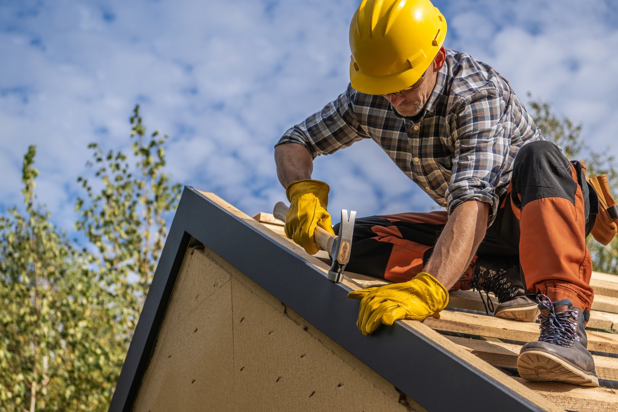 Roofer wearing a yellow hard hat and gloves, installing roofing materials on a sloped roof, surrounded by trees and blue sky, illustrating professional roofing services related to commercial and residential projects.