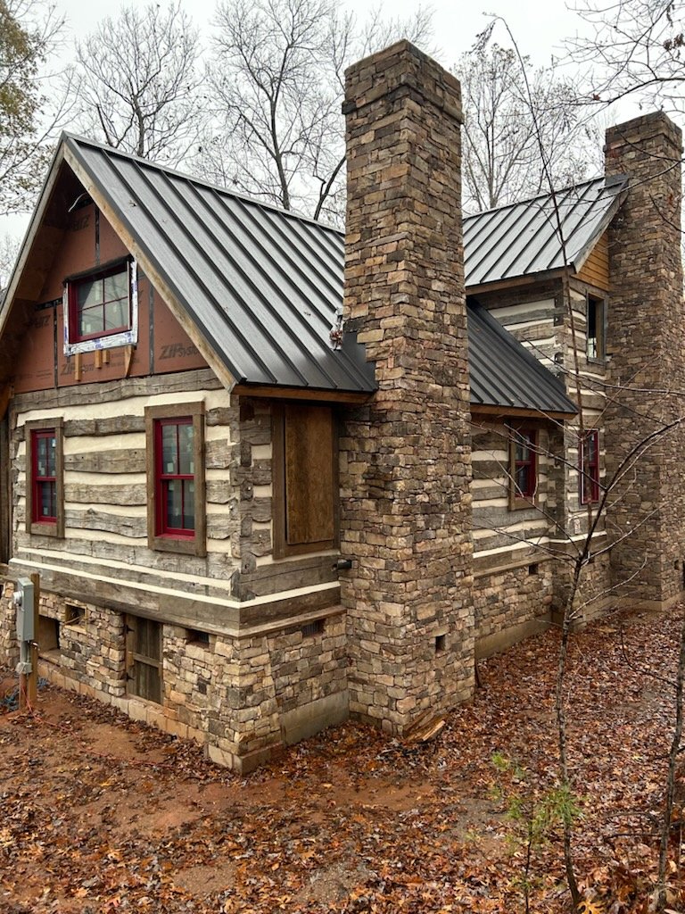 Historic log cabin with stone chimney and metal roofing, surrounded by autumn leaves, illustrating unique challenges in roof replacement for historical homes.
